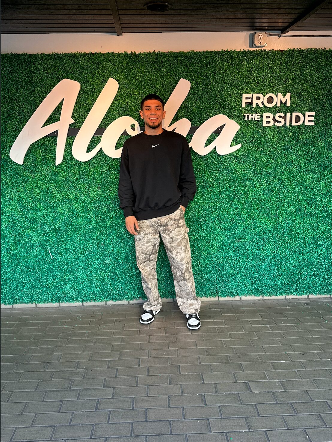 UNKODUKE in front of an Aloha sign on a green hedge wall